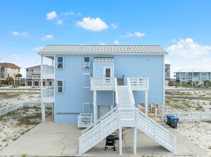 Une maison de plage à deux étages, avec un escalier blanc menant à un balcon, entourée de sable et d’autres maisons de plage.