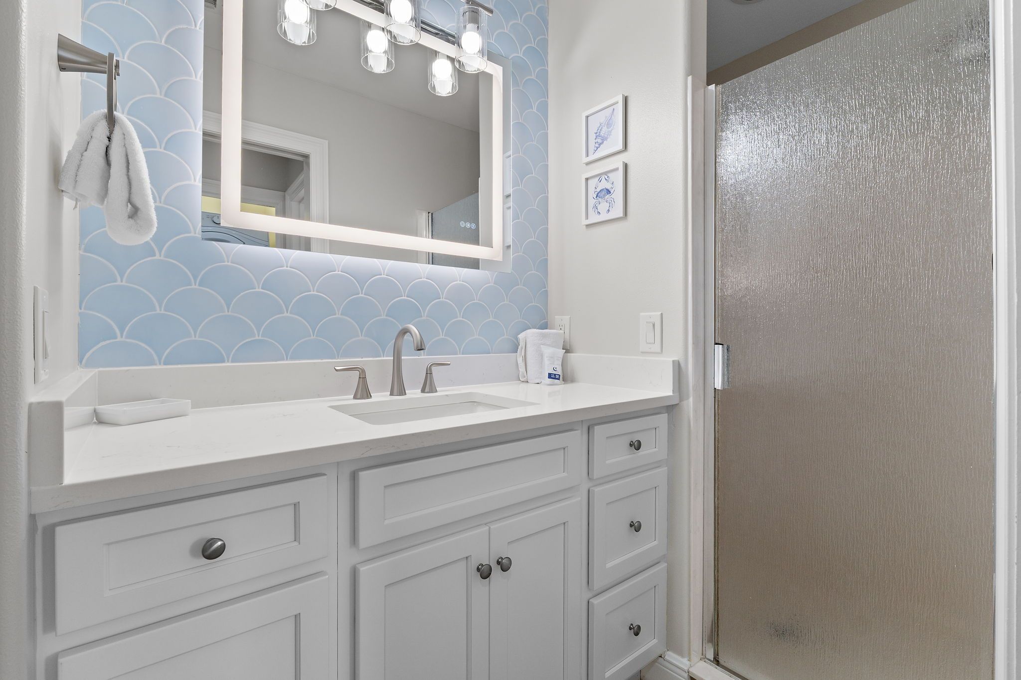 A bathroom with a white vanity, a mirror with lights, a towel rack, and a door with a textured surface.