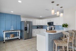 A modern kitchen with white cabinets, a stainless steel refrigerator, a microwave, and a coffee maker. There is a white island with a sink and a wooden chair. - Jake's Sand Dollar: Newly Remodeled + Walk to Navarre Beach 🌊 (Navarre Beach)