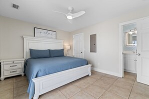 Une chambre à coucher comprenant un lit, une table de chevet, une lampe fixée au mur, un ventilateur de plafond et une porte donnant sur une salle de bain.