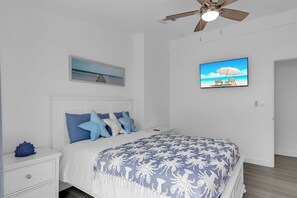 Ein Schlafzimmer mit Bett, Nachttisch, Deckenventilator, einem an der Wand montierten Fernseher und einem gerahmten Bild von einer Strandansicht.