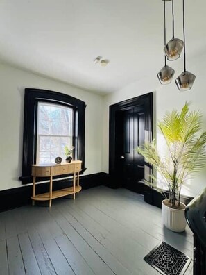Ein Zimmer mit Holzboden, einem Fenster mit schwarzem Rahmen, einer schwarzen Tür, einem Blumentopf und einem Holzkonsole mit einer Vase.