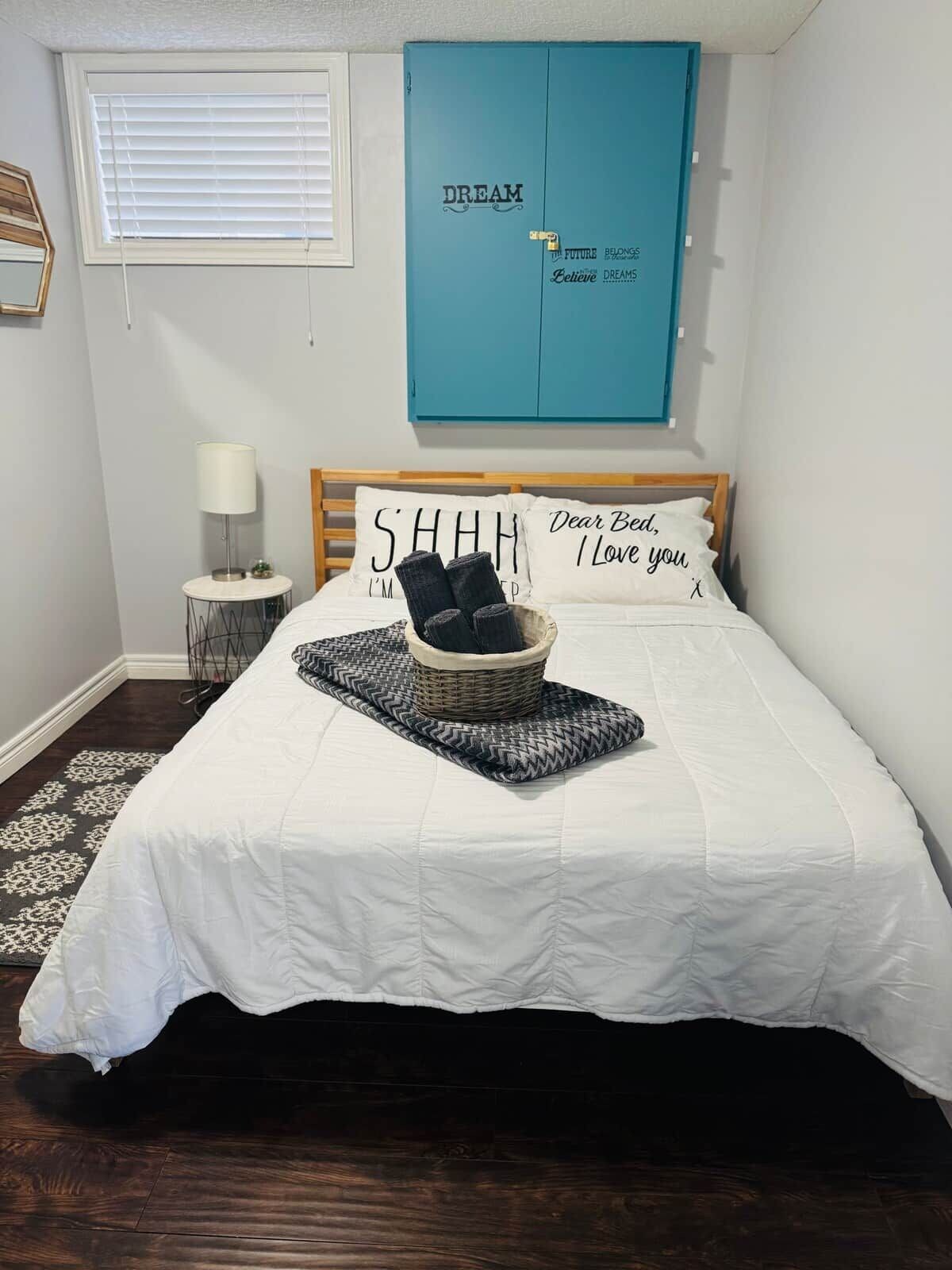 A bedroom with a bed, a basket of items, a lamp, and a wall cabinet.