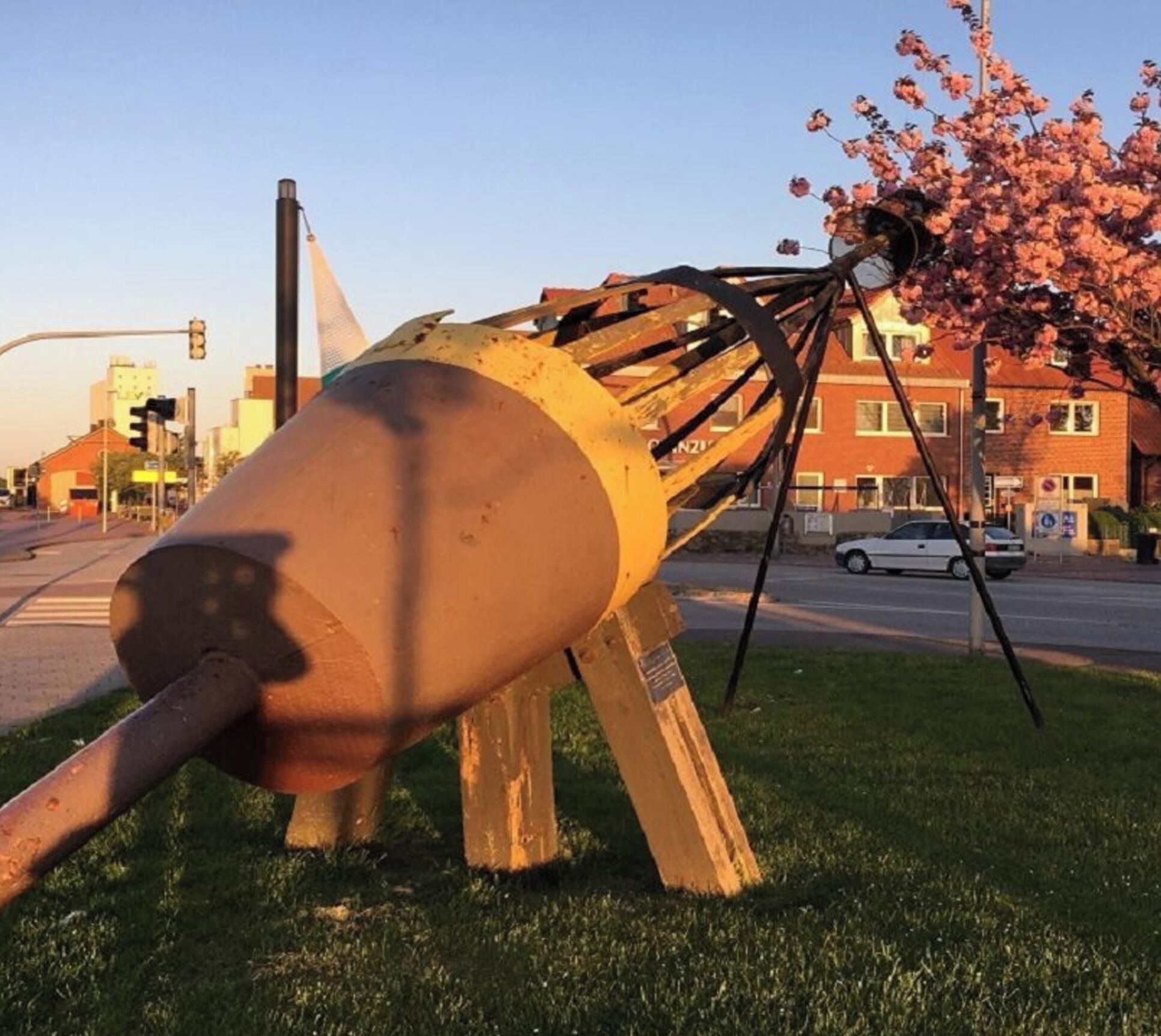 A large, metallic sculpture with a cylindrical base and a complex structure on top, situated in a grassy area with a street and buildings in the background.