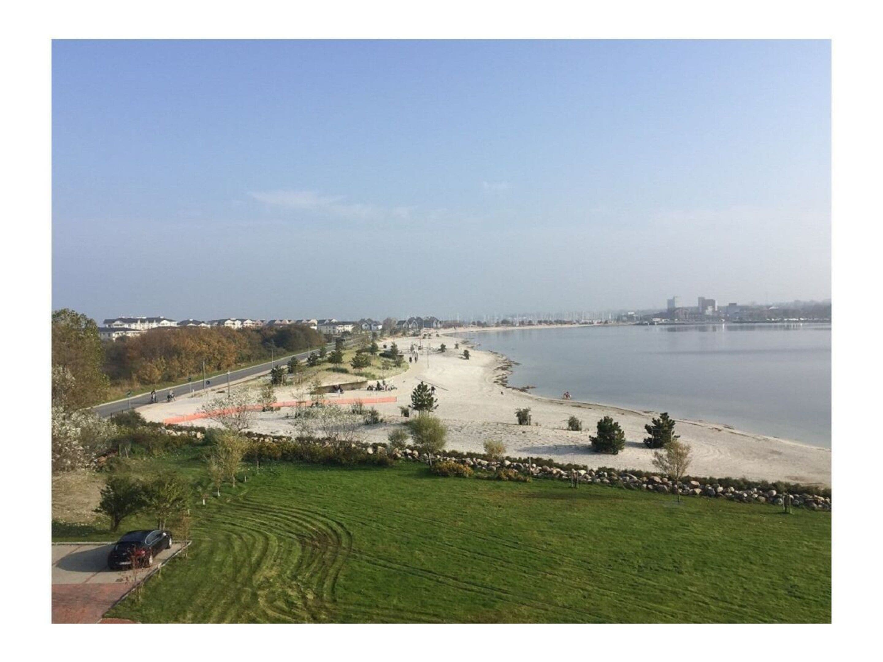 Un paysage côtier avec une plage de sable, une zone herbeuse et une route menant à la silhouette d'une ville au loin.