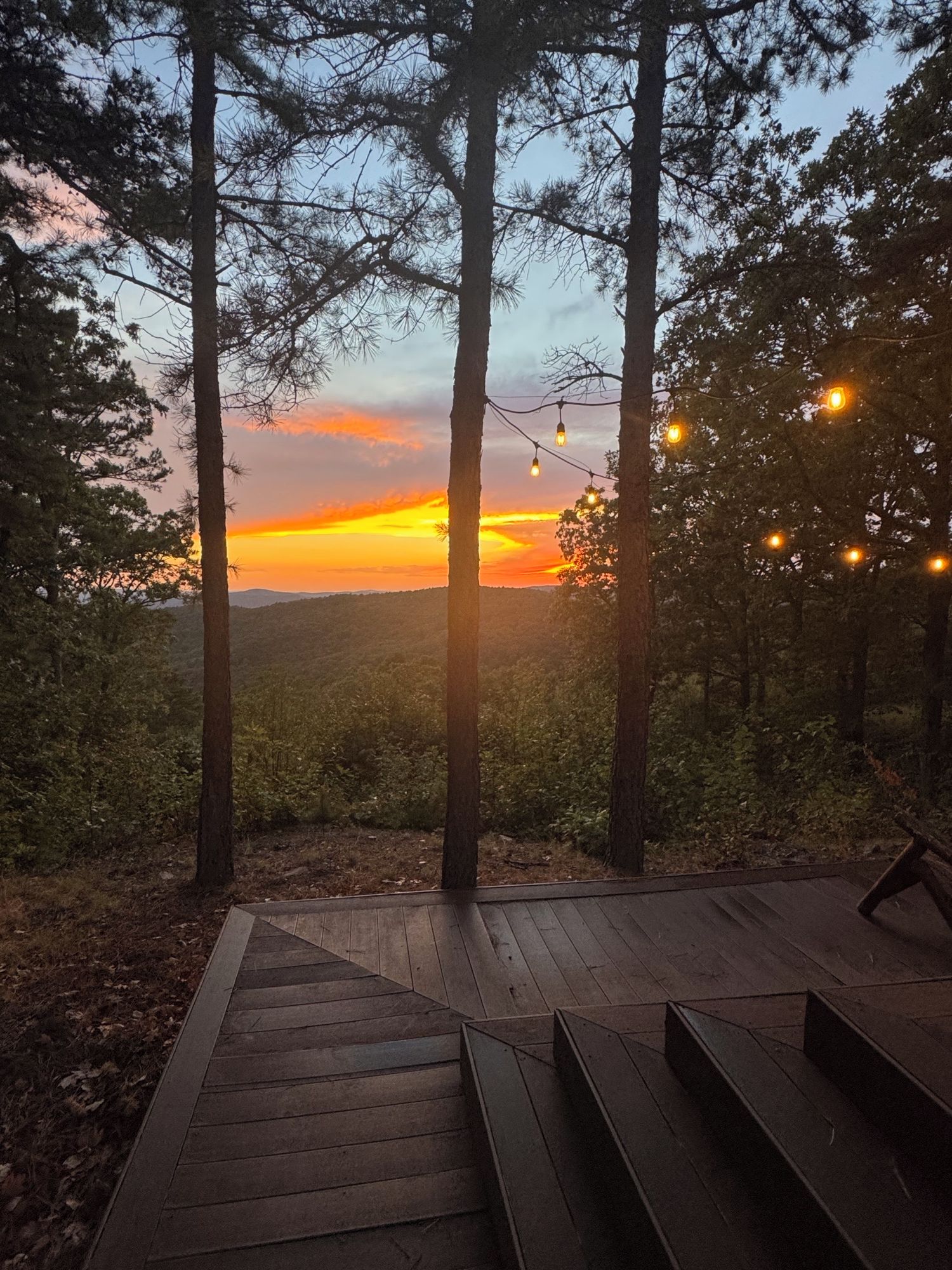 Et træterrasse med trapper ned til et skovområde, med udsigt over en solnedgang.