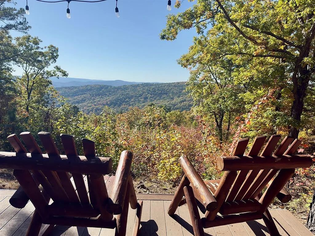 Deux fauteuils Adirondack en bois sont placés sur une terrasse en bois, d’où l’on peut admirer un paysage boisé et, au loin, une chaîne de montagnes.