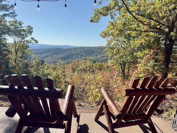 Two wooden Adirondack chairs on a wooden deck overlooking a forested landscape with a distant mountain range.