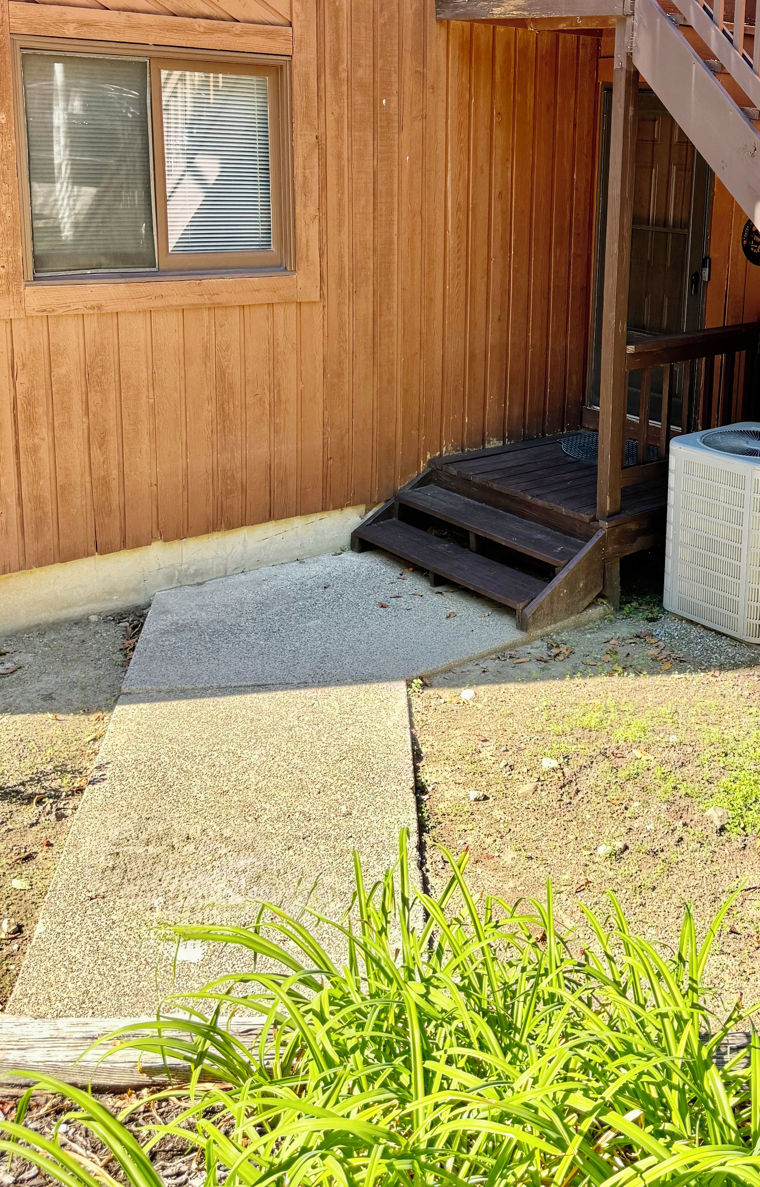 En träbyggnad med ett fönster, en trappa som leder upp till en veranda och en gångbana av betong.