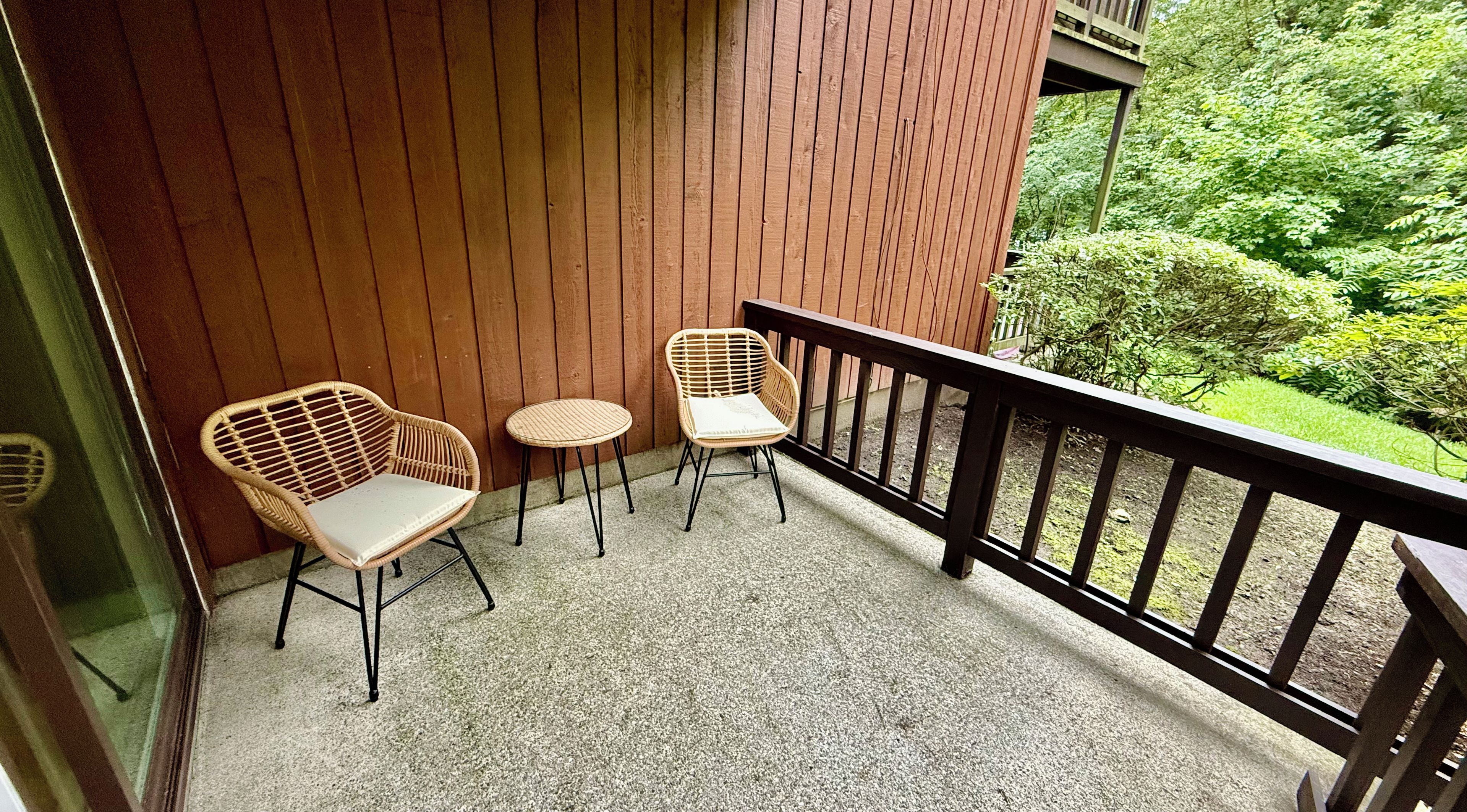 Un balcon avec deux fauteuils en osier et une petite table ronde, une balustrade en bois et une vue sur la verdure.