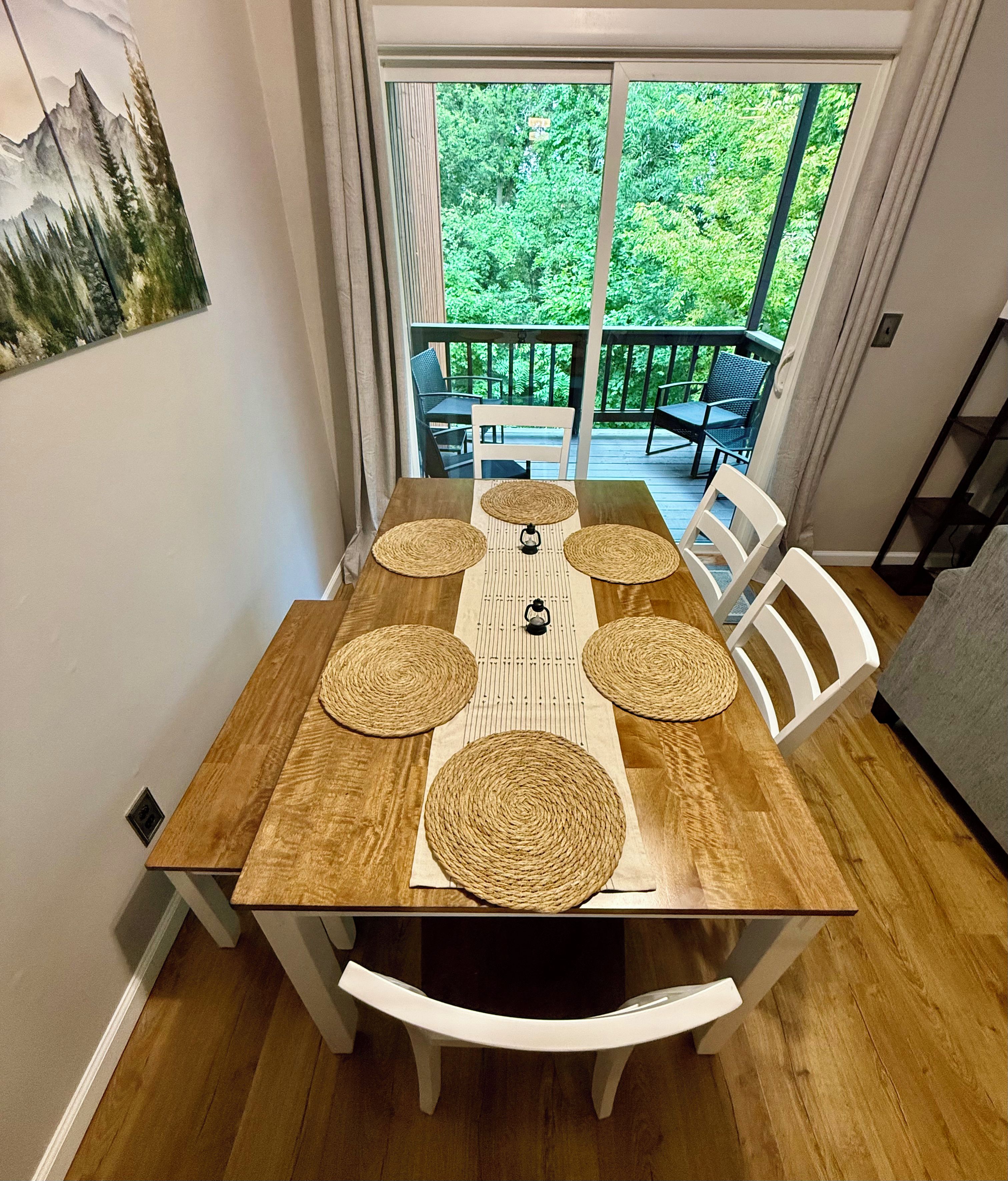 A dining table with woven placemats and white chairs, set on a wooden floor with a view of greenery through a glass door.