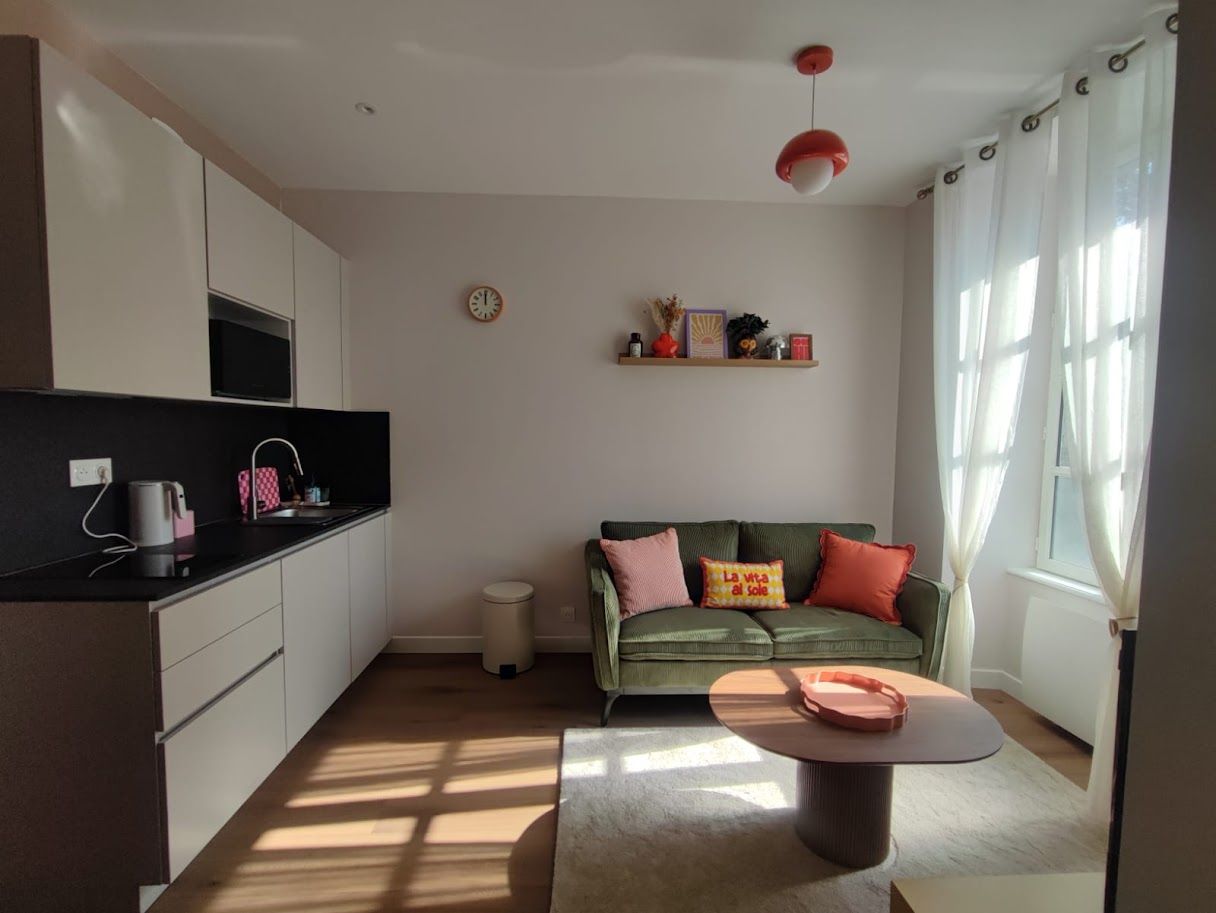 A modern living space with a kitchen, a green sofa with cushions, a coffee table, and a window with curtains.