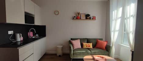 A modern living space with a kitchen, a green sofa with cushions, a coffee table, and a window with curtains.