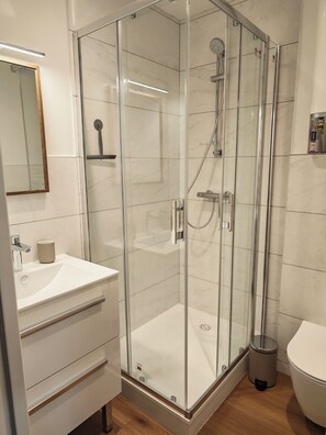 Un baño moderno con ducha envuelta en vidrio, un lavabo blanco con una taza, y un inodoro.