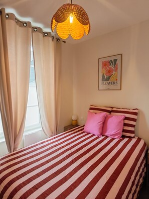 Una cama con rayas rojas y blancas, una lámpara colgante, un cuadro con flores y cortinas.