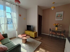 Una sala de estar con sofá, una mesa de centro, un televisor sobre un mueble amarillo, una mesa de comedor con sillas y una ventana con cortinas.