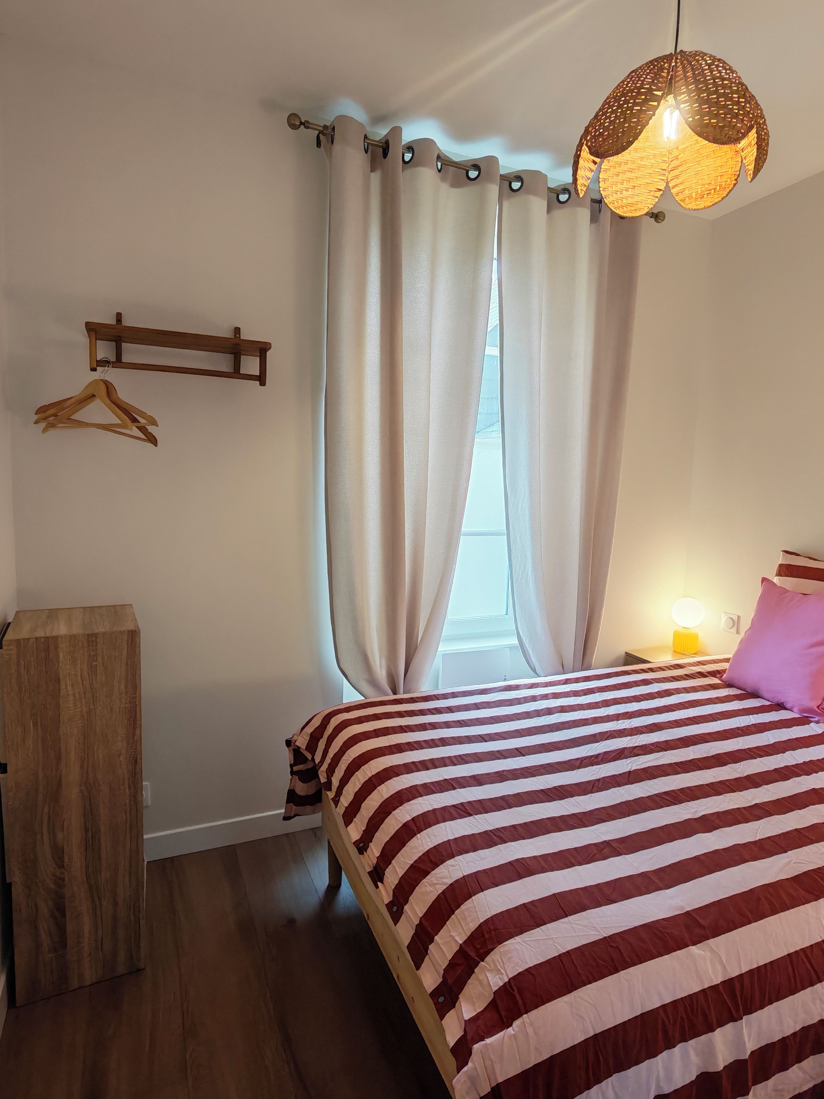 Een slaapkamer met een bed, gordijnen, een hanglamp en een houten dressoir.