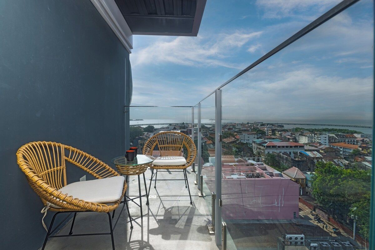 A balcony with wicker chairs and a glass railing overlooking a cityscape.