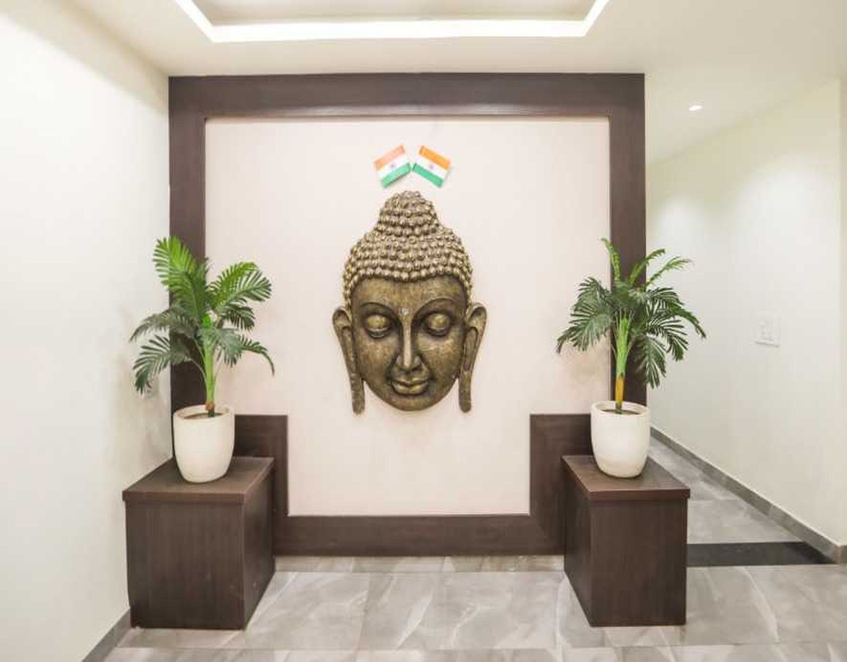 Escultura de cabeça de Buda fixada na parede, com uma fita tricolor acima, e dois vasos de plantas em pedestais escuros em um corredor.