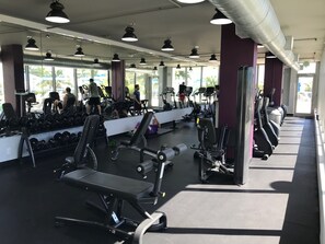 Une salle de sport équipée de divers appareils d’exercice, de miroirs et d’un mur vitré qui laisse entrer la lumière naturelle.
