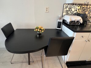 Un coin repas avec une table noire, deux chaises, un vase de fleurs, un petit présentoir à menus et un comptoir de cuisine équipé d’appareils électroménagers.