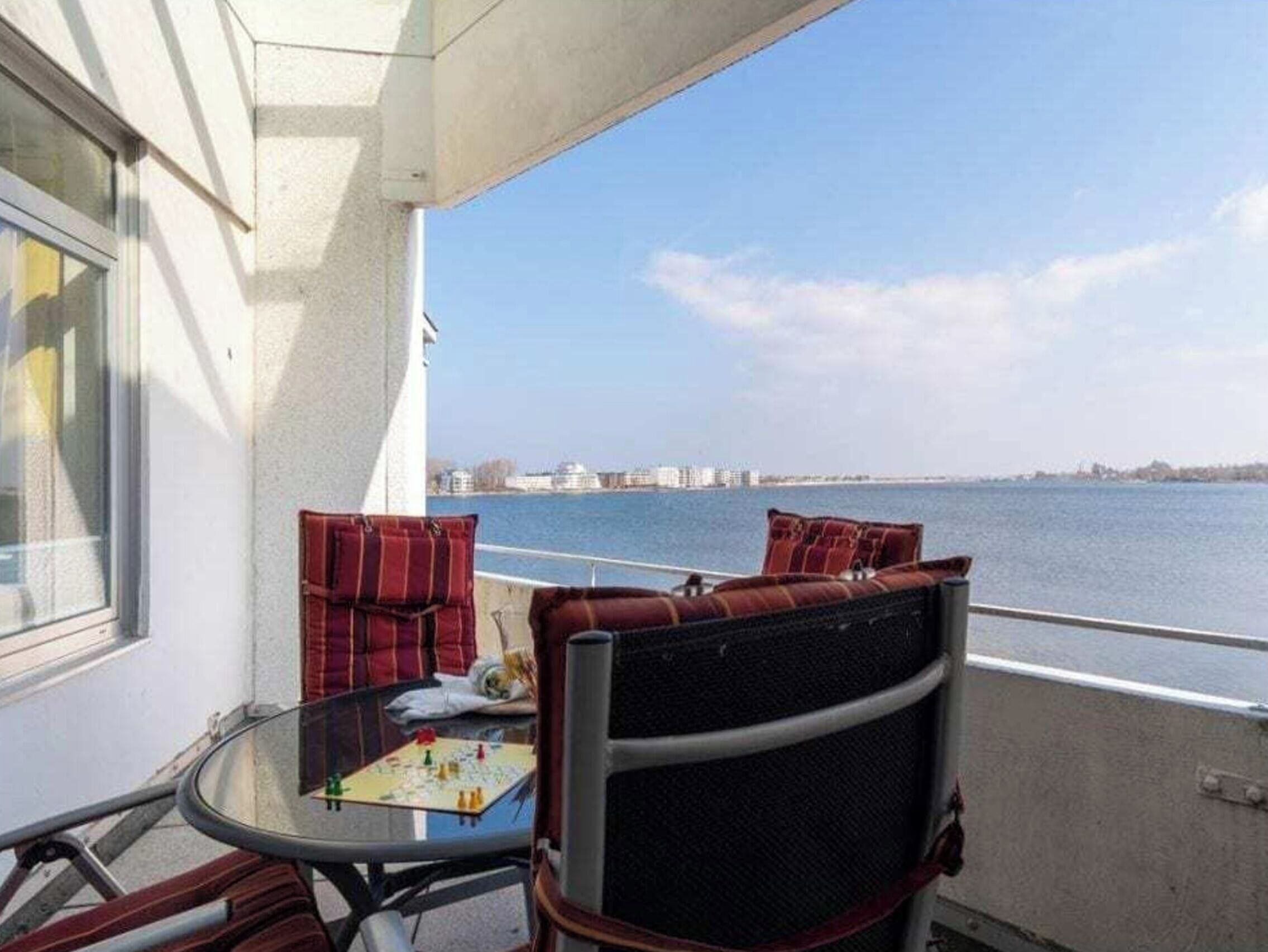 Ein Balkon mit Tisch und Stühlen, mit Blick auf ein Gewässer und entfernte Gebäude.