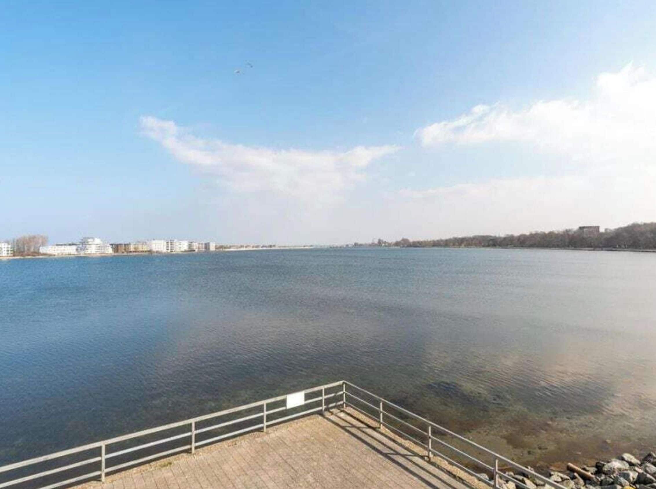 A calm body of water with a clear sky, a wooden dock, and distant buildings.