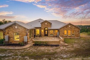 A two-story stone house with a metal roof, a front porch, and a well-maintained lawn. - Solterra Hilltop | Pool | Grill + Endless Views! (Fredericksburg)
