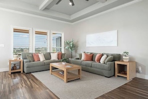 A living room with a sectional sofa, a coffee table, and a large window with blinds. - Solterra Hilltop | Pool | Grill + Endless Views! (Fredericksburg)