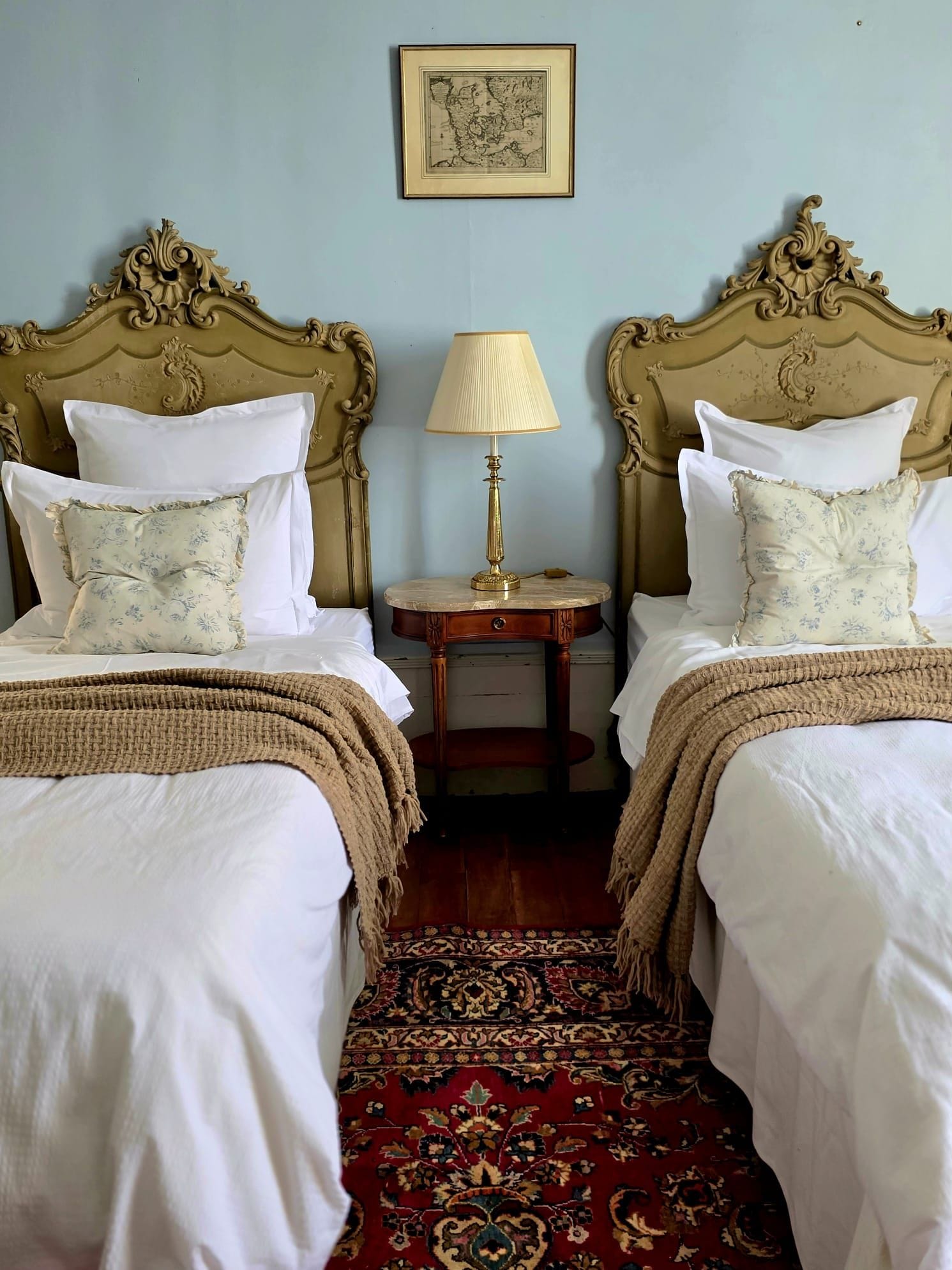 A room with two beds, a small table with a lamp, and a patterned rug.