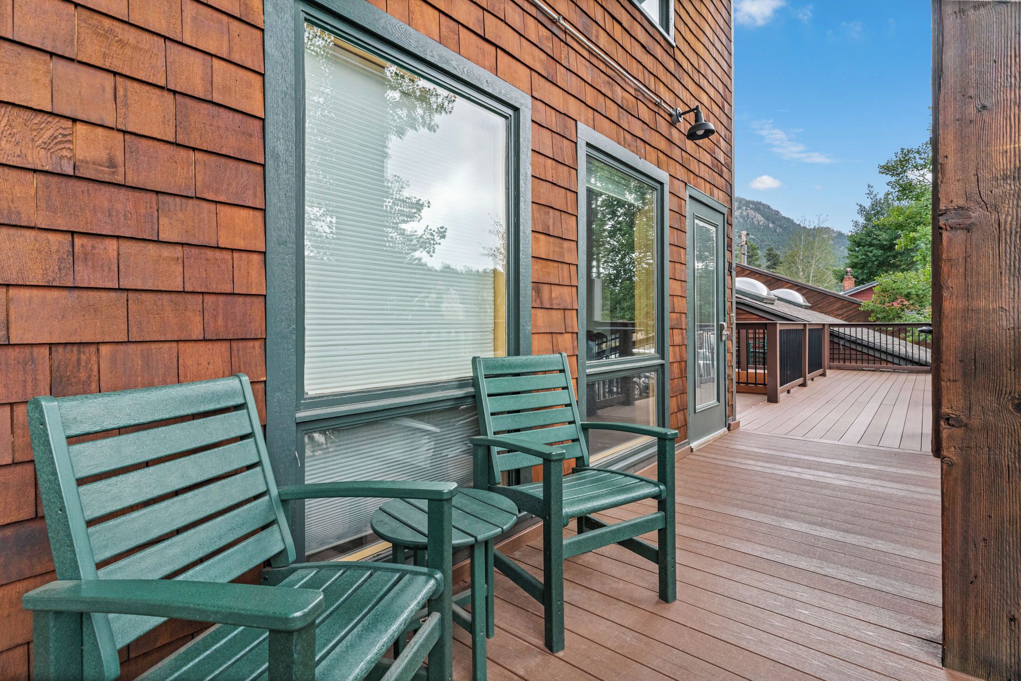 Une terrasse en bois avec deux chaises vertes et une table, un bâtiment en bois doté de grandes fenêtres et un ciel dégagé.