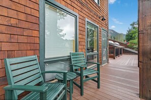 A wooden deck with two green chairs and a table, a wooden building with large windows, and a clear sky.