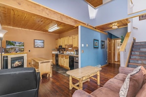 A cozy living room with a fireplace, wooden furniture, and a kitchen area with wooden cabinets and appliances.