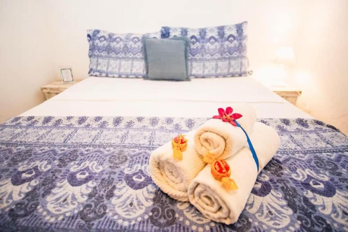 A bed with a patterned bedspread, a blue pillow, and rolled towels with decorative flowers.