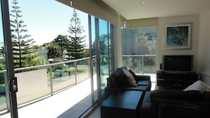 Una sala de estar moderna con sofá, mesa de centro y televisor. Amplias ventanas ofrecen vista a árboles y una carretera.