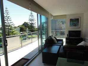 A modern living room with a sofa, a coffee table, and a TV. Large windows offer a view of trees and a road. - 507 at The Waves (Cowes)