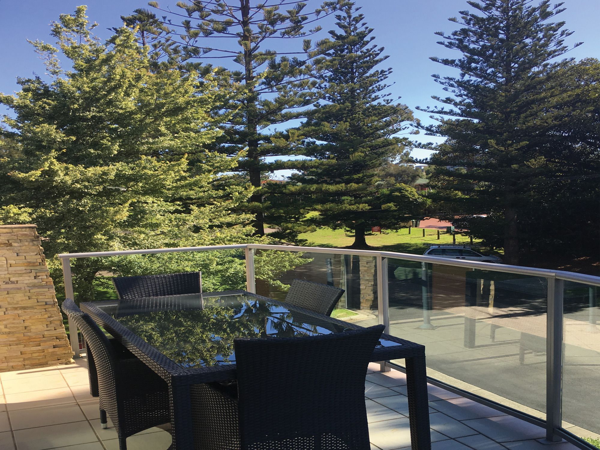 An outdoor dining area with a glass balustrade, wicker furniture, and a view of trees and a building in the background.