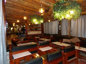 Interior de restaurante com mesas e bancos de madeira, plantas penduradas e um ambiente acolhedor.