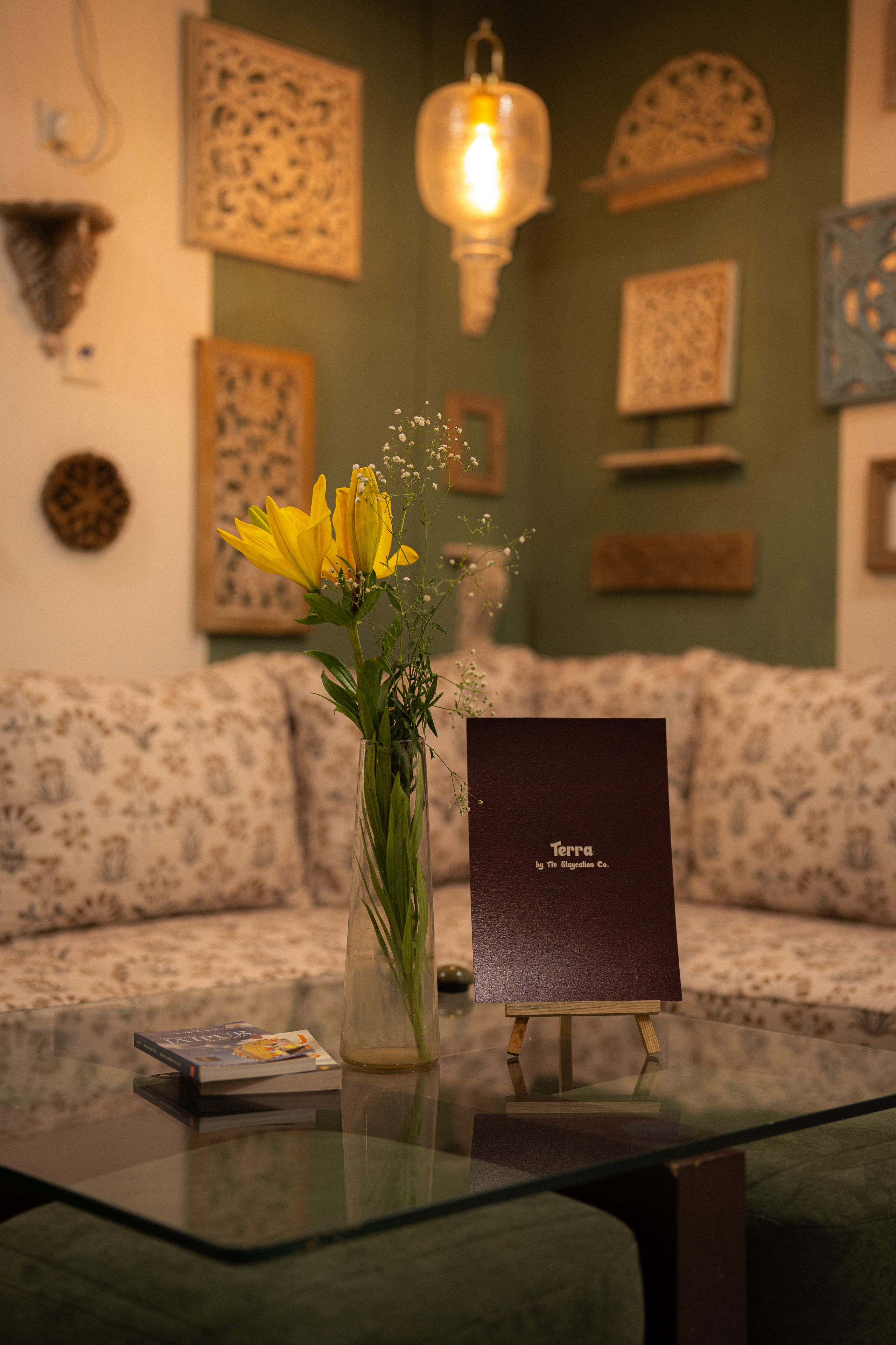 A room with a glass coffee table, a vase with yellow flowers, a framed book titled 'Terra by The Slaycation Co.', and a patterned sofa.