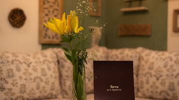 A room with a glass coffee table, a vase with yellow flowers, a framed book titled 'Terra by The Slaycation Co.', and a patterned sofa.