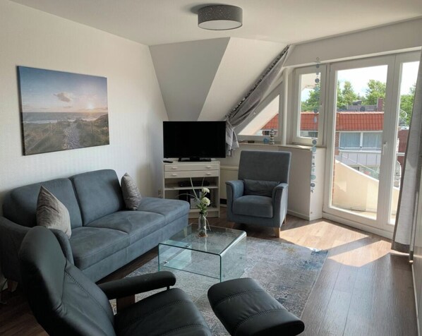 Ein modernes Wohnzimmer mit einer Couch, Sesseln, einem Glastisch, einem Flachbildfernseher und einem Landschaftsbild.