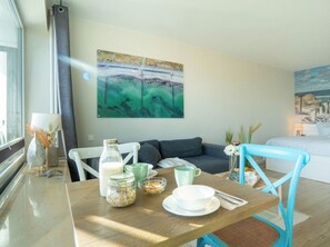 A room with a dining table set with a glass jar, a cup, and a bowl. There is a sofa and a bed in the background. - Steinwarder 7, Apartment "strandbude 33" (Heiligenhafen)