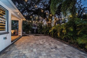 Une terrasse dallée, agrémentée de guirlandes lumineuses, entourée d’une végétation luxuriante et bordée d’un bâtiment à la façade blanche.
