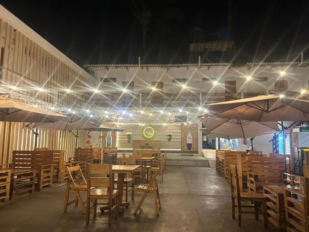 An outdoor dining area with wooden tables and chairs, umbrellas, and a building with a corrugated metal facade in the background.