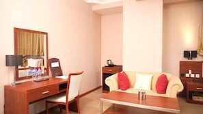 Habitación de hotel con escritorio de madera, una silla, un sofá con cojines rojo y blanco, una mesita de centro y un armario pequeño.