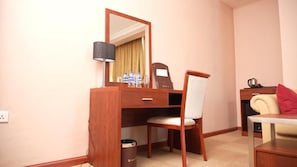 Habitación de hotel con un escritorio de madera, una silla, un espejo, una lámpara, un cubo de basura y una mesita con un cojín rojo.