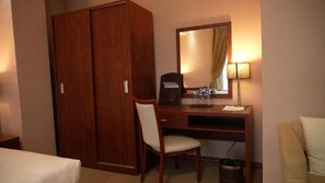 Een hotelkamer met een houten bureau, een stoel, een bed, een kledingkast, een spiegel, een lamp en een prullenbak.