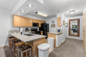 A modern kitchen with wooden cabinets, a black refrigerator, a microwave, and a stove. There are bar stools and a trash can. - Snowy Owl Lodge | Walk to Slopes & Shared Hot Tub (Breckenridge)