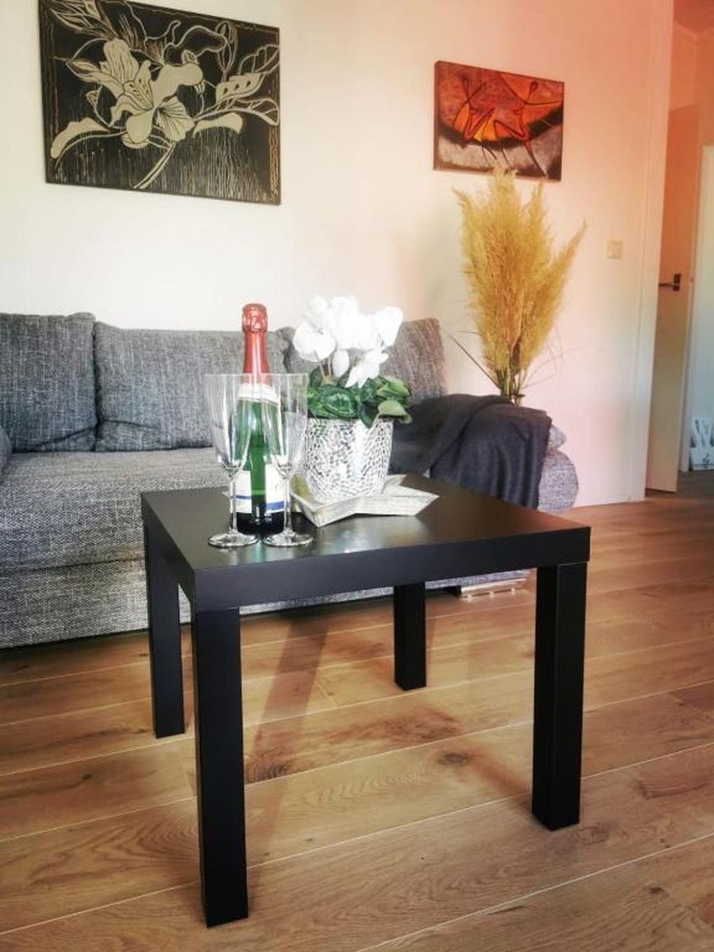 Un salon avec une table basse noire, un canapé gris et un mur orné de deux œuvres d’art.