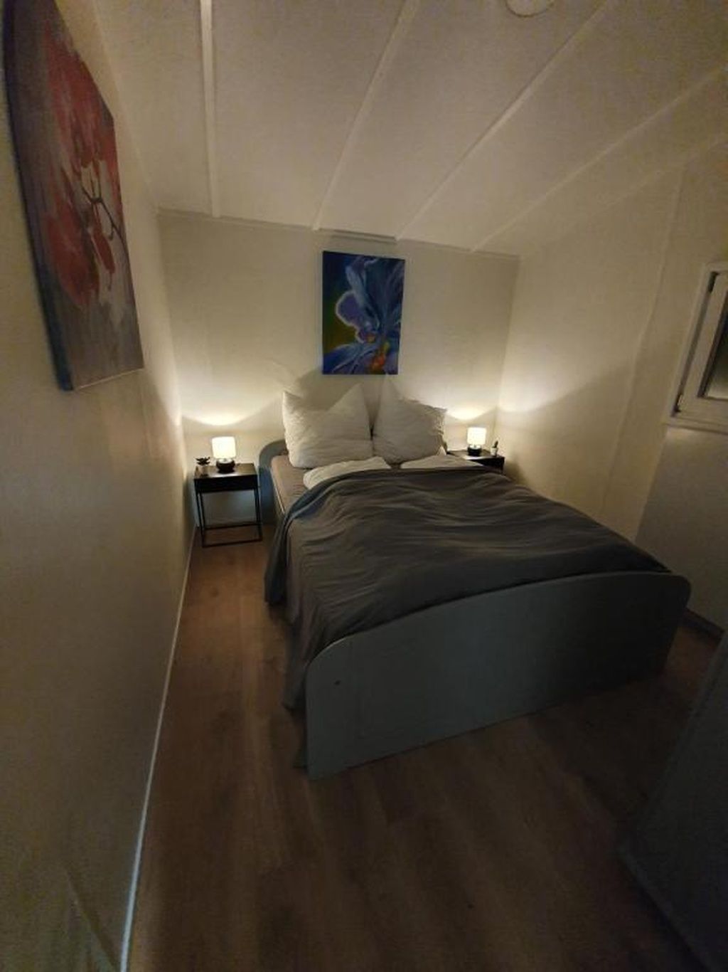 A bedroom with a bed, two bedside tables, a nightstand, a painting on the wall, and a window.